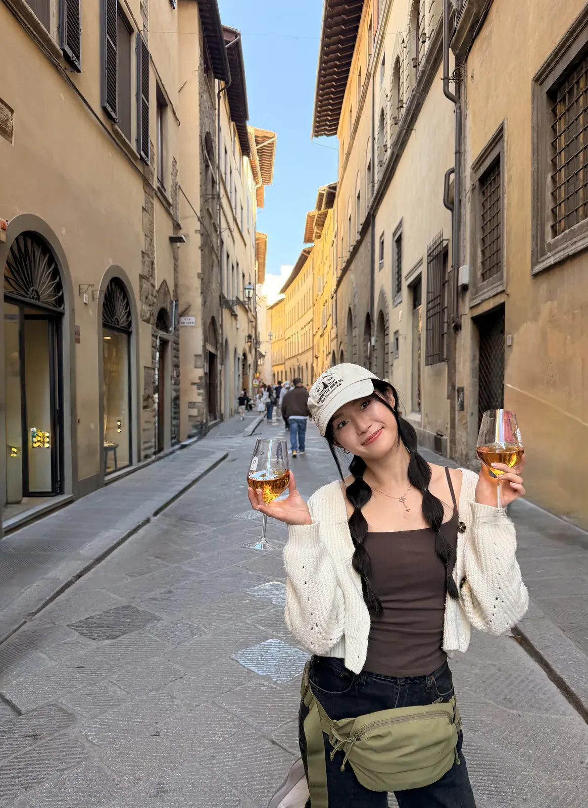 A traveler holding up two glasses of wine on a narrow stone-paved Oltrarno street, with shuttered buildings on both sides and evening light on the facades at the far end