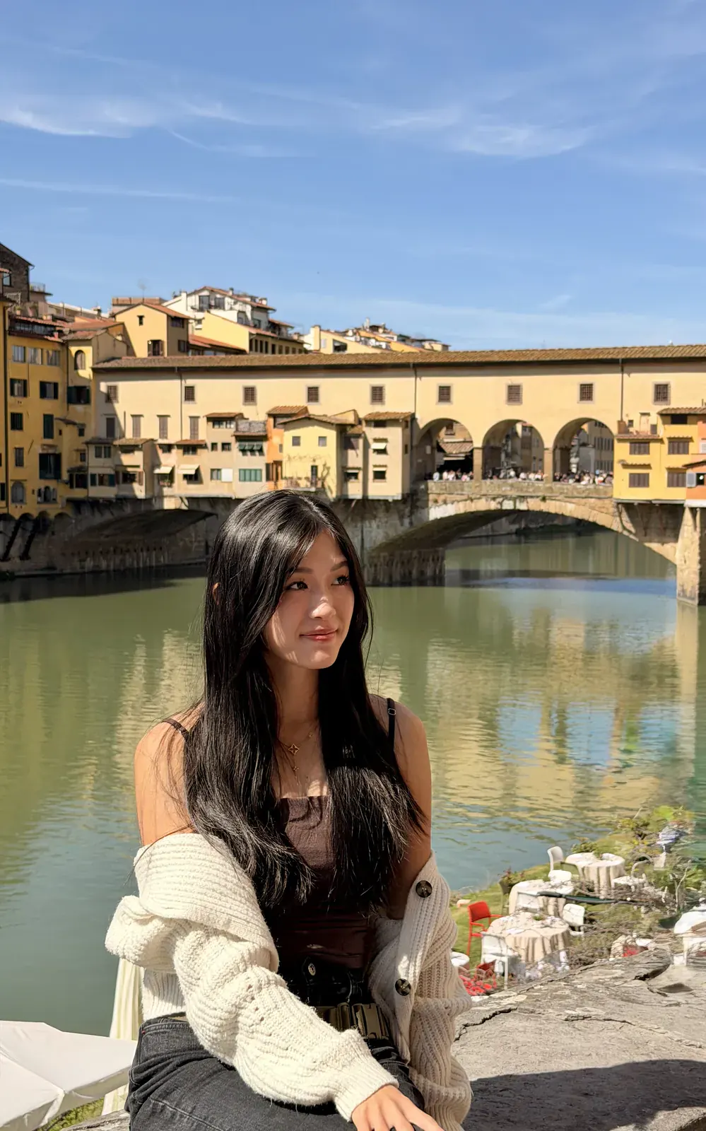 A traveler sitting on the bank of the Arno in front of Ponte Vecchio, with the shops built along the bridge and the covered corridor running above them