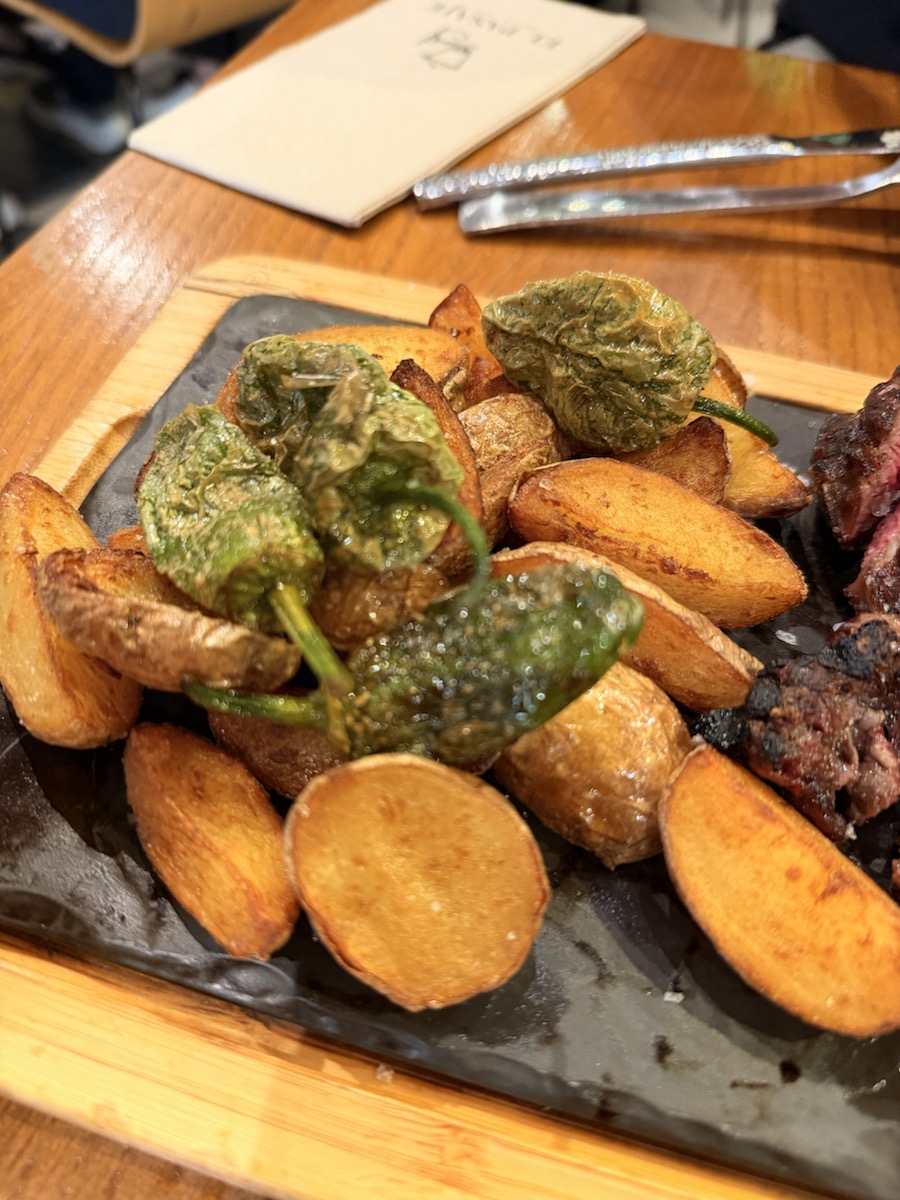 Fried potatoes topped with blistered green peppers beside grilled meat at a tapas bar